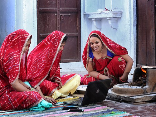 Three traditional Indian young married women working in traditional kitchen on laptop. Using technology in rural households. Women cook food and learn technology. Rural women using Laptop.